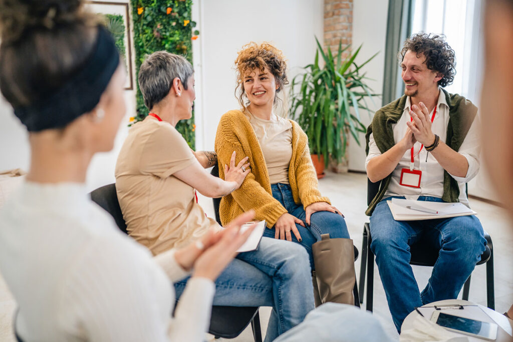 A group of people sitting in a circle with two therapists and a woman in the center discussing and providing emotional support for her The focus is on the woman but everyone is engaged in conversation