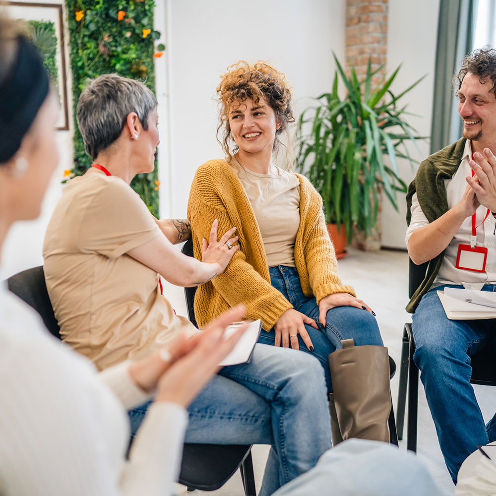 A group of people sitting in a circle with two therapists and a woman in the center discussing and providing emotional support for her The focus is on the woman but everyone is engaged in conversation