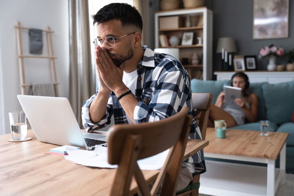 Young nervous concentrated man working on laptop calculating home finances while woman lying on sofa reading and surfing the net. Domestic life of freelancer worker and his girlfriend.